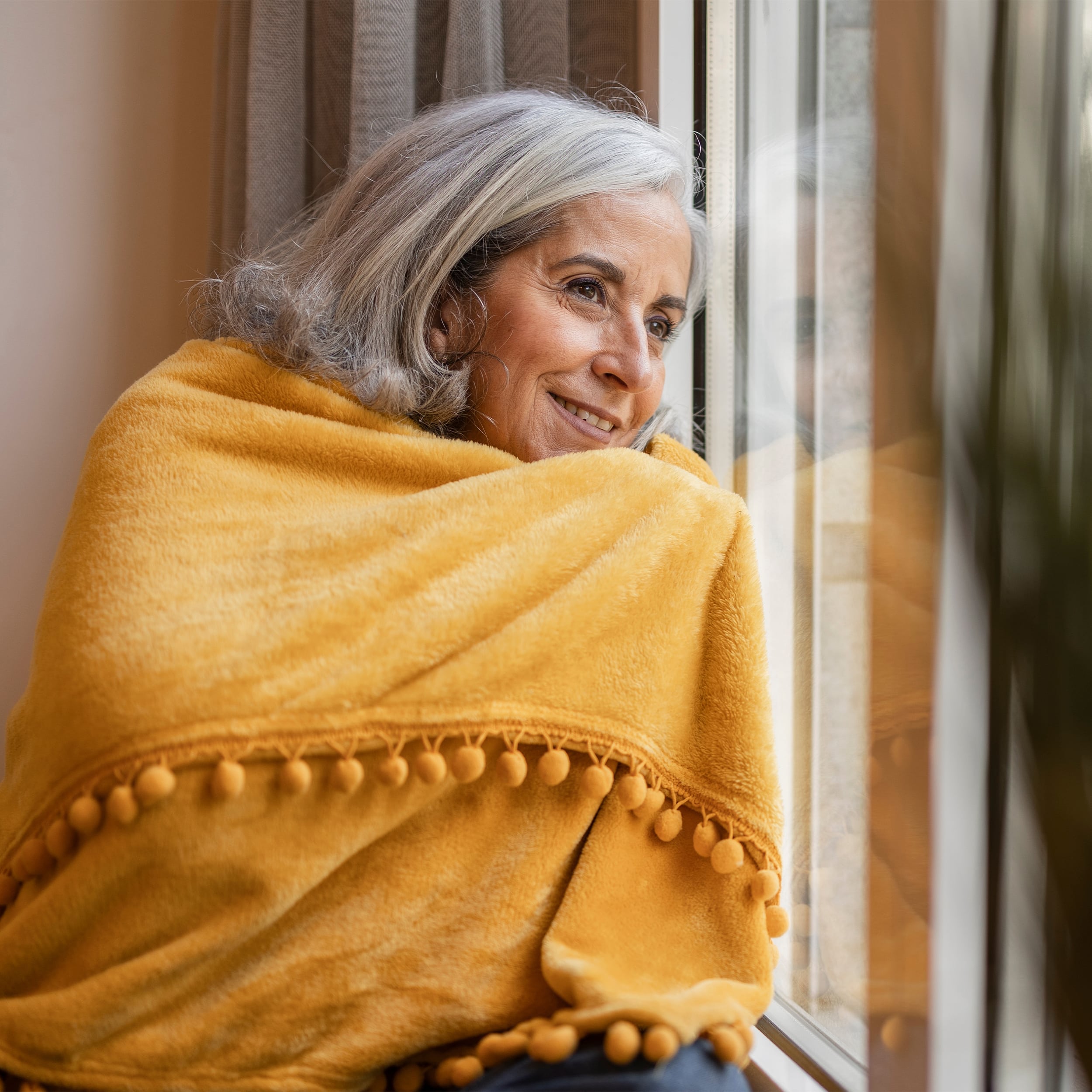 Eine ältere Dame mit weissen Haare hat sich in eine gelbe Decke gehüllt. Sie lehnt gegen ein Fenster und schaut nach draussen.