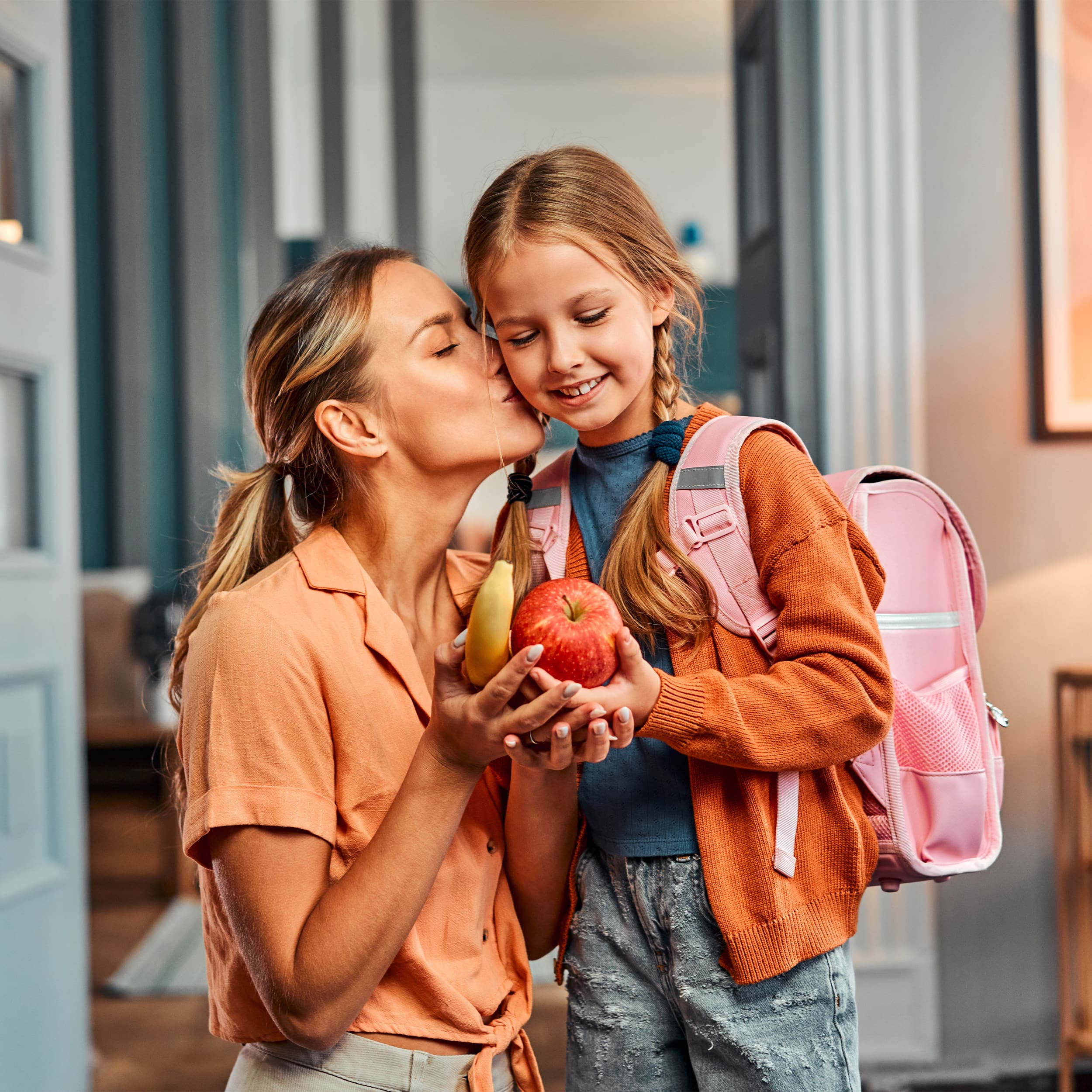 Ein kleines Mädchen steht mit einem Schulrucksack und einem Apfel und einer Banane in der Hand in einem Hausflur. Ihre Mutter kniet an ihrer rechten Seite am Boden und gibt ihr gerade einen Kuss auf die Wange.